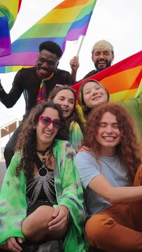 Vertical. Group of young LGBT poeple smiling celebrating together the gay pride day with a rainbow flag. Diverse friends looking at camera and having fun on a rights parade. Equality and freedom