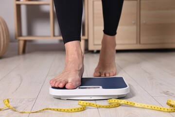 Woman stepping on floor scale and measuring tape at home, closeup. Weight control