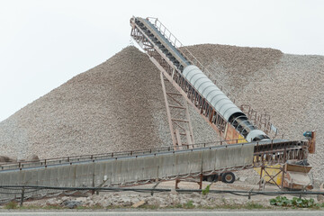 A stockpile of bulk limestone mined in an above ground quarry. The beige pelleted crushed sedimentary rock of magnesium carbonate and calcium in raw form is piled in a mound. The background is white.