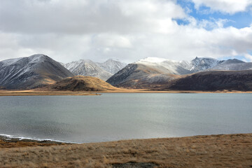 The gentle shores of a wide lake with ice along the edges lies in the endless steppe at the foot of snow-capped mountains on a cloudy autumn day.