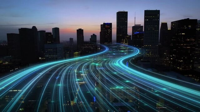 American city at dusk with curving light trails, glowing blue digital connectivity lines animation. Aerial dolly forward towards dark skyscrapers at night.