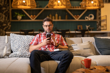 Handsome cheerful man indoors at home on sofa using mobile phone. Attractive caucasian man resting...