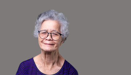 Portrait of an elderly Asian woman looking at the camera with a smile while standing on a gray...