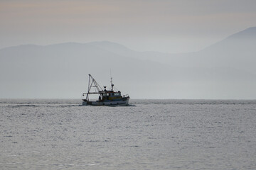 A small commercial fishing boat heads out to sea in Tasman Bay, New Zealand almost silhouetted against hazy morning sun.