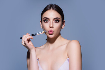 Girl hold blush blusher apply powder visage isolated over studio background. Young woman powdering cheeks. Makeup brush. Female model gets blush powder on the cheekbones.