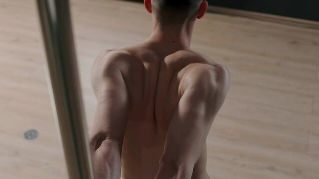 Back view of shirtless young male pole dancer with hands behind his back wrapped around pole doing stretching exercise on his back during indoor training in dark dance studio