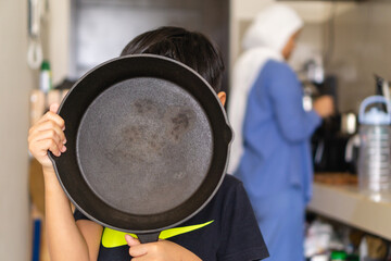kids holding cast iron skillet with a background of his mom cooking in the kitchen, cocept of...