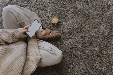 Woman sitting on fluffy carpet with cup of coffee and using mobile phone with blank screen. Cozy...