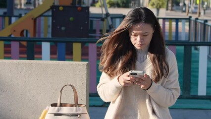 A Chinese woman at the kindergarten in a Sapnish park. Young mother  uses her smartphone. Family oriented activities and modern technology, childhood education. 