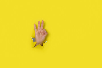 Hand making a sign of OK, coming out of the hole in a torn yellow paper background.