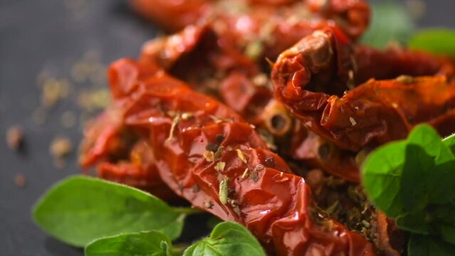 Sun dried tomatoes with aromatic herbs, oregano, marjoram leaf. Rotating on black stone background, close up. Italian cuisine, healthy vegan food, Mediterranean diet. Dry tomato