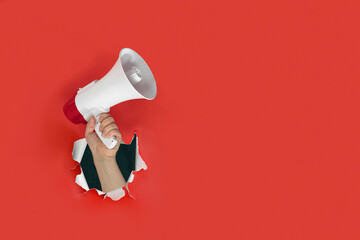 Hand holding loudspeaker through a hole in paper red background. Announcement, advertising, public...