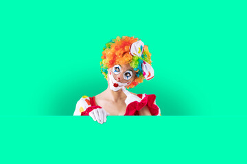 Thoughtful look female clown peeking out from behind sign blank template mock up for advertising, posters, greetings, invitations. Entertainer as Joker in a suit and wig, with clown whiteface makeup.