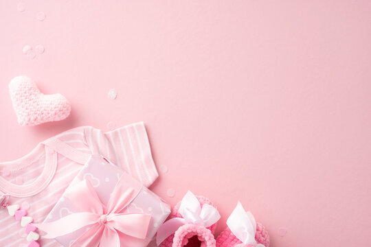 Flat lay of pink baby shower items including baby clothes, gift box, crocheted heart, and baby shoes on pink background