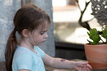 A little sad girl with a ponytail stands by a pot with an indoor flower on the terrace