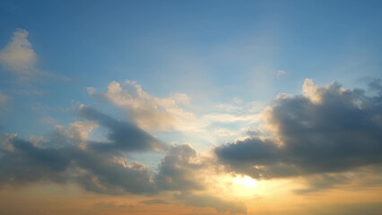 A sunset with the sun partially obscured by clouds. The sky transitions from soft blue to warm orange near the horizon, with clouds reflecting the sun's golden light. Cloud background.
