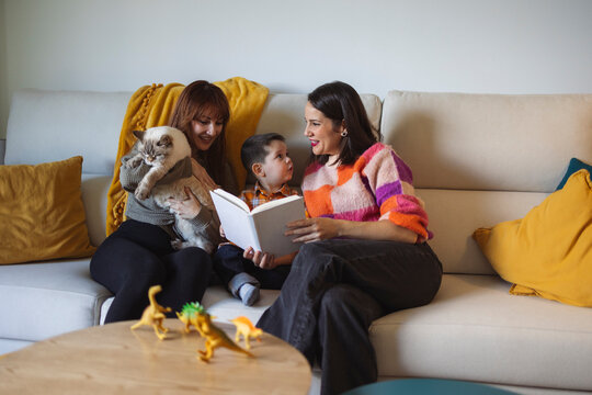 Happy mother reading book to son with woman holding cat on sofa at home