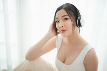Naklejka premium A beautiful young woman with long dark hair and a white sleeveless top wearing headphones, enjoying music in a bright room. She looks serene and content, with a subtle smile and soft lighting.