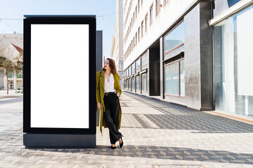 Fashionable businesswoman leaning on blank billboard at footpath in city