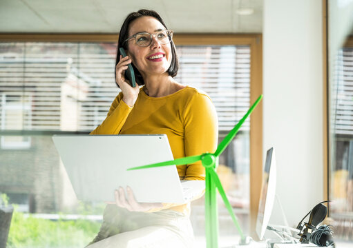 Smiling businesswoman talking on mobile phone using laptop sitting by wind turbine model in office