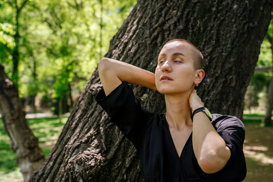 Woman with shaved head leaning on tree trunk