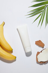 Cosmetic advertising photo with an unlabeled tube displayed between some coconut pieces and bananas on the white flat, a green coconut leaf decorated in the top corner. Overhead shot photo, copy space © Tuan  Nguyen 