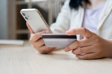 A close-up image of an Asian woman sits at a table indoors, holding her smartphone and a credit card