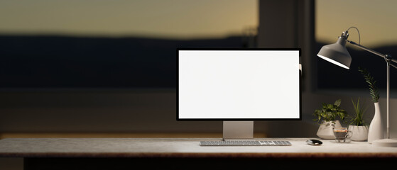 A computer desk in a modern, dark room in the evening features a white-screen computer mockup.
