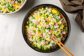 Cantonese Fried Rice with egg, green pea, ham steak, spring onion, ribe long rice in a dark bowl on a white table directly above.
