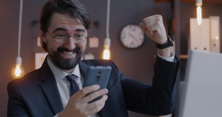Overjoyed Middle Eastern entrepreneur expressing happiness looking at smartphone screen celebrating success in office at night. Business and good news concept. - Powered by Adobe