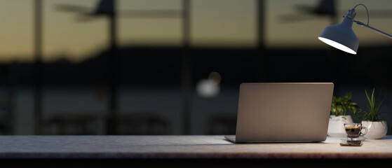 A modern, dark office workspace at night features a laptop computer and dim light from a table lamp.