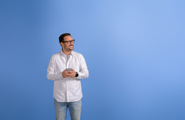 Happy male entrepreneur with smart phone looking away and thinking business ideas on blue background