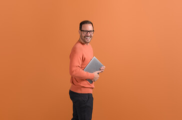 Portrait of cheerful young man holding laptop and laughing at camera on isolated orange background