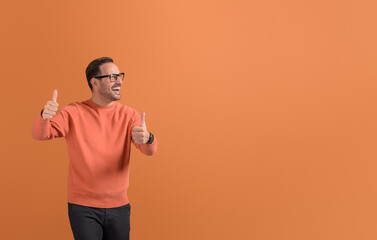 Successful young businessman showing thumbs up sign and looking away cheerfully on orange background