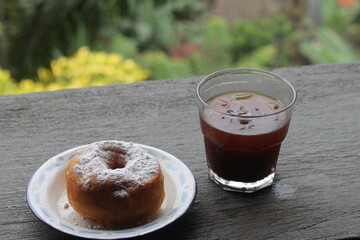A sugar powdered donut sits on a white enamel plate next to a glass of iced black coffee. Selective focus