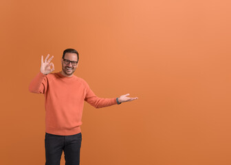 Portrait of smiling young businessman showing empty palm and OK sign on isolated orange background