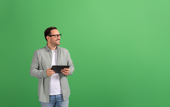 Successful businessman with digital tablet smiling and looking away thoughtfully on green background - Powered by Adobe