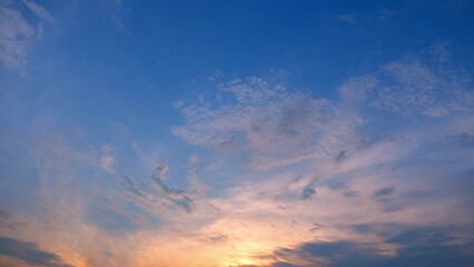 A serene sunset with a sky adorned with wispy and scattered clouds. The lower part of the sky is illuminated with soft hues of orange and pink, while the upper part transitions into a clear blue.
