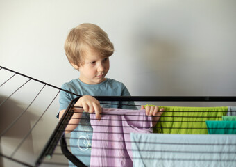 Cute 5 year old boy hanging wet clothes on a metal drying rack, helping his mother, help around the...