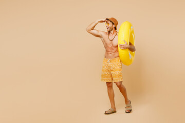 Full body fun happy young man wear yellow shorts swimsuit hat relax rest near hotel pool with rubber ring hand at forehead look far away isolated on plain beige background. Summer sea sun tan concept.