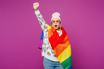 Young lesbian woman wrapped in rainbow flag in pink animal clothes raise up hands clench fist scream shout isolated on plain purple background studio portrait Pride day June month love LGBTQ concept