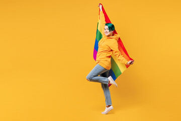 Full body side view young lesbian woman with dyed green hair wears hoody casual clothes hold rainbow flag behind herself isolated on plain yellow orange background. LGBT diversity lifestyle concept.