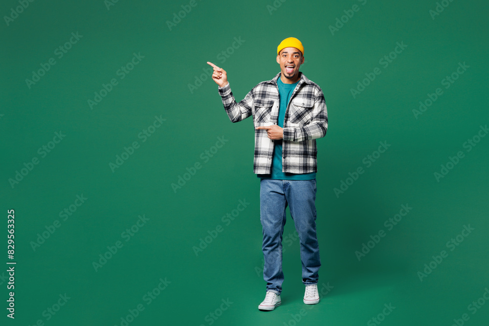 Wall mural Full body surprised young man of African American ethnicity he wear shirt blue t-shirt yellow hat point index finer aside on area isolated on plain green background studio portrait. Lifestyle concept. - Wall murals