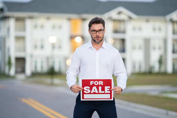 Handsome real estate agent holding banner house for sale. House with land and insurance. Business man real estate agent in business suit presenting the house for sale. Land agent trying to sell house.
