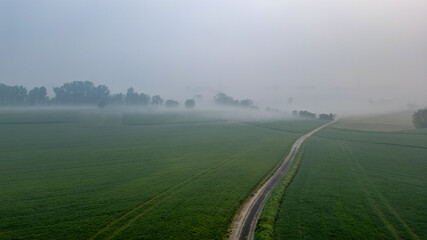 This aerial photograph captures a serene misty morning over expansive farmland. The image features a winding path that stretches through lush green fields, gradually fading into the mist. Trees dot