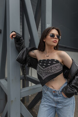 Fashion beautiful young woman model with stylish sunglasses in fashionable clothes with a bandana top with a leather jacket and jeans stands near a metal construction outdoors