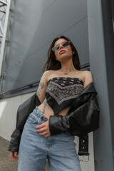 Glamorous fashion urban woman with cool sunglasses in trendy clothes with bandana top and leather jacket with jeans stands in the city