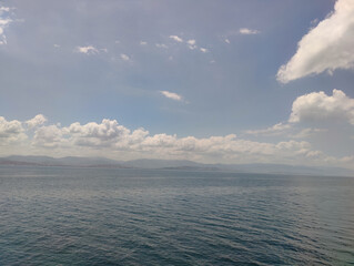 A serene marine scene with deep blue-green water, small waves, a motorboat, larger vessels, and a clear, slightly cloudy sky.