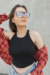 Beautiful fashion student woman with fashionable glasses in stylish clothes with a tank top and shirt against the background of a concrete wall
