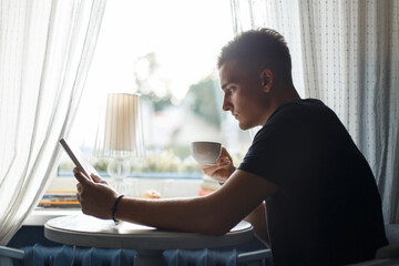 young man sits in a cafe by the window, drinks coffee and reads a book